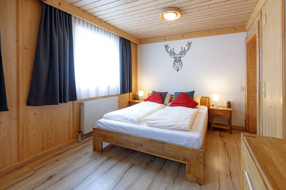 A wooden-paneled bedroom with a bed, two bedside tables, a radiator, and a wall-mounted deer head decoration.