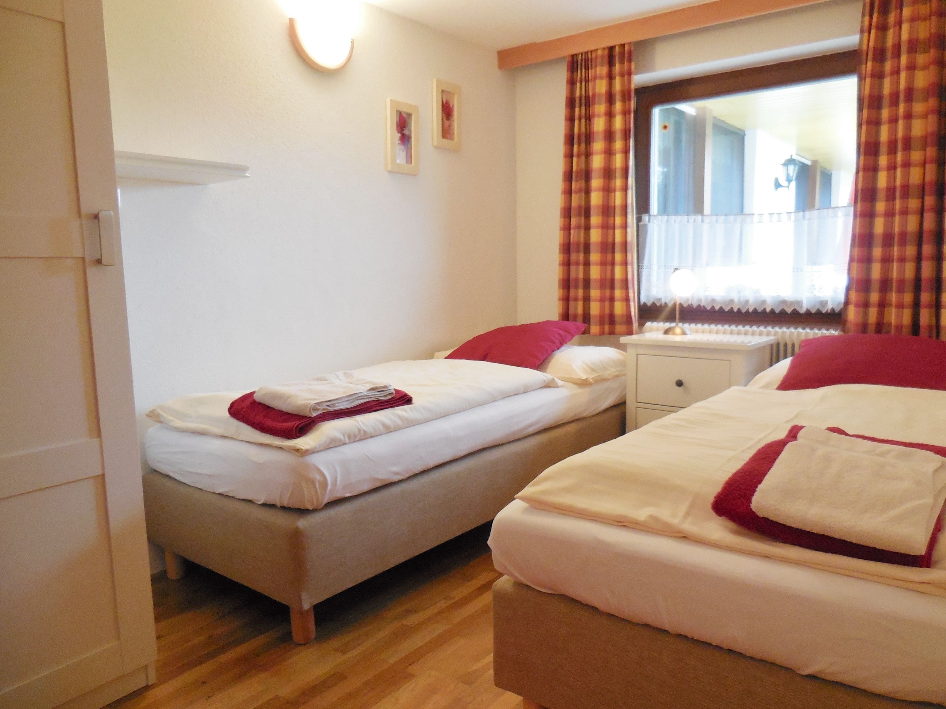 A hotel room with two single beds, a small bedside table, a nightstand, a window with curtains, and a wall-mounted light fixture.