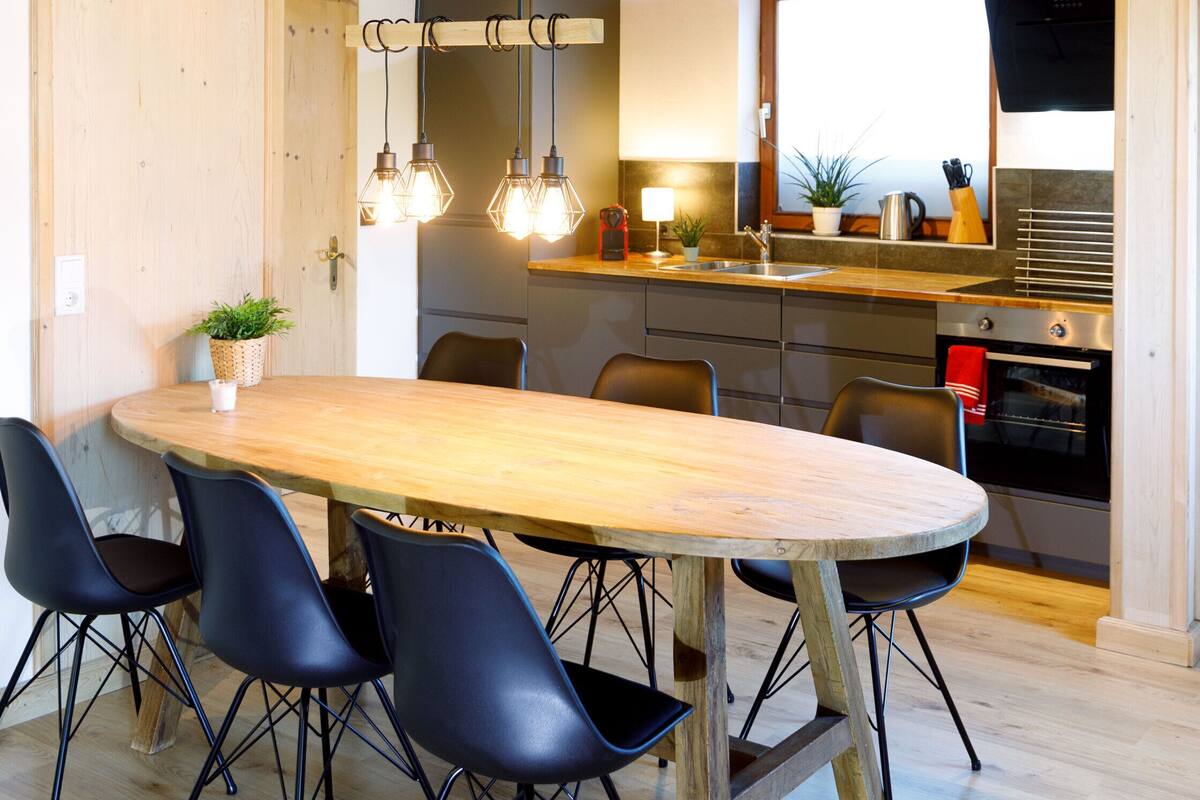 A modern kitchen with a wooden table and chairs, a hanging light fixture, and a built-in oven.