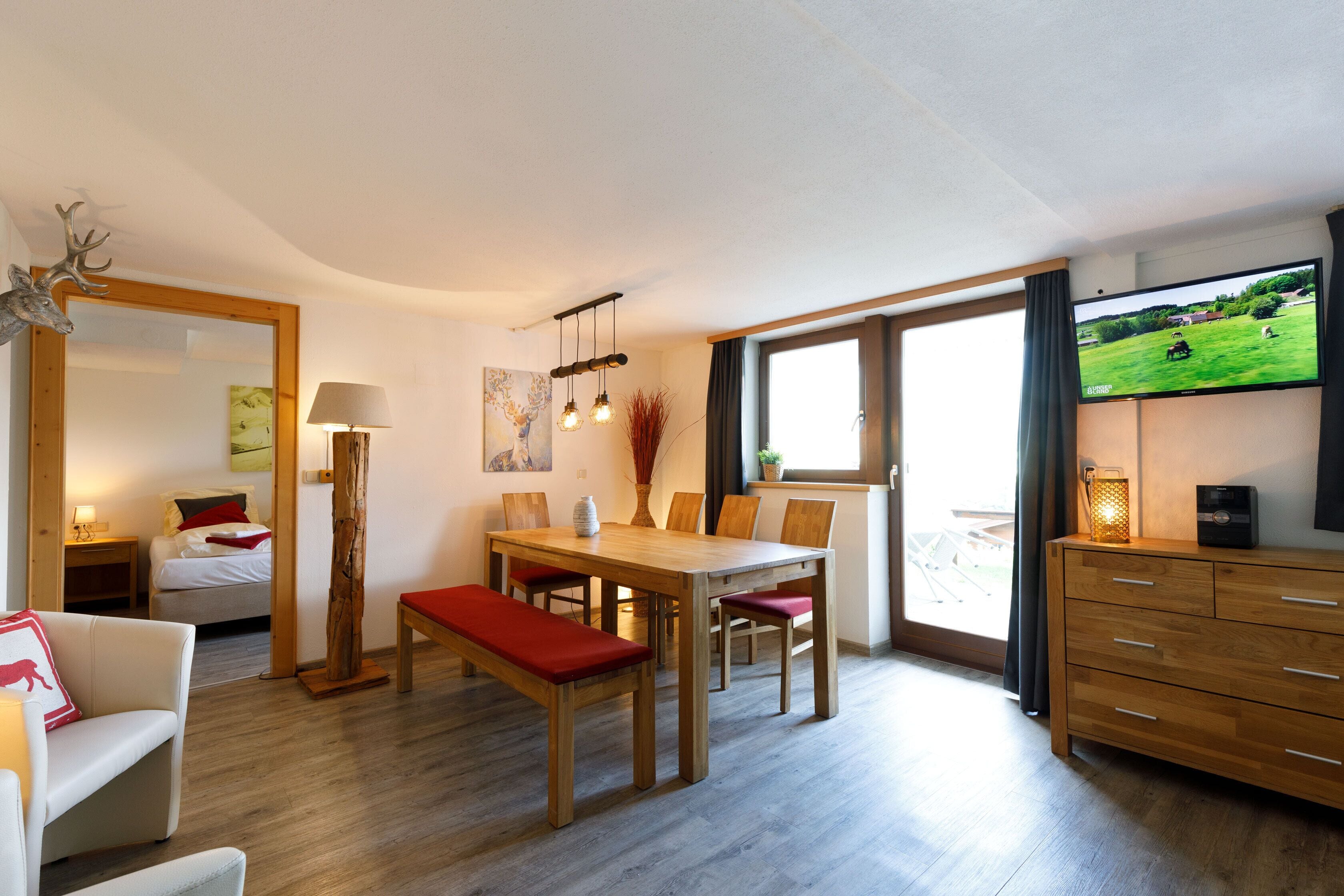 Una sala de estar moderna con una mesa de comedor de madera, un banco con cojines rojos y un televisor montado en la pared.