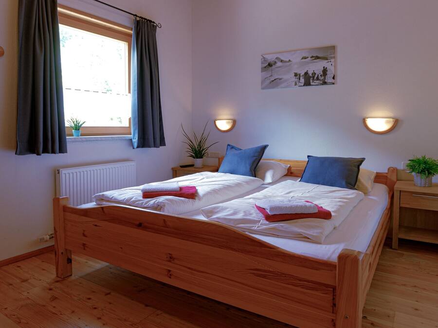 Una cama con estructura de madera, dos almohadas, una mesita de noche con lámpara y una ventana con cortinas.