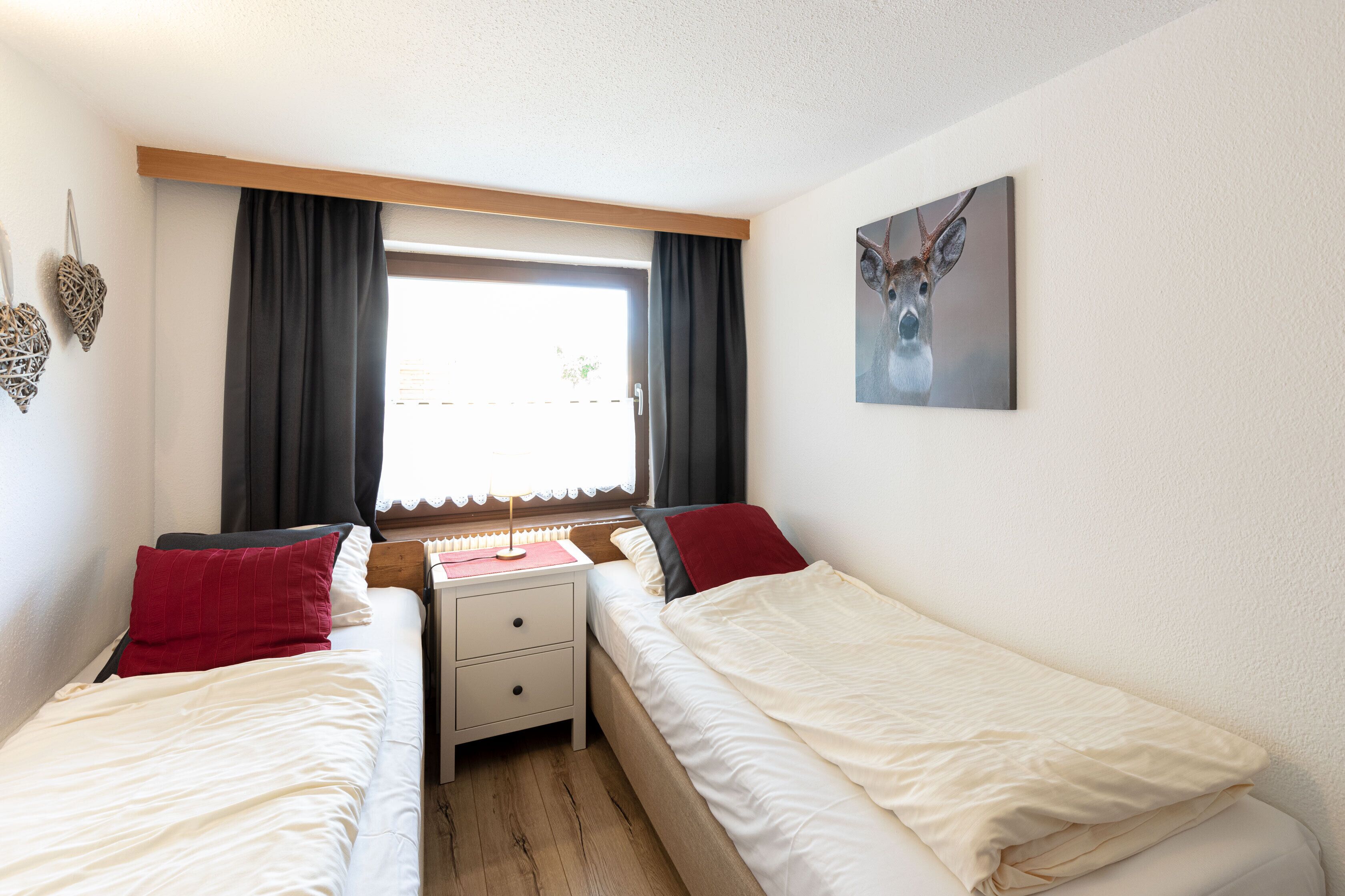 Une chambre avec deux lits, une table de chevet, une fenêtre avec des rideaux et un tableau représentant un cerf accroché au mur.