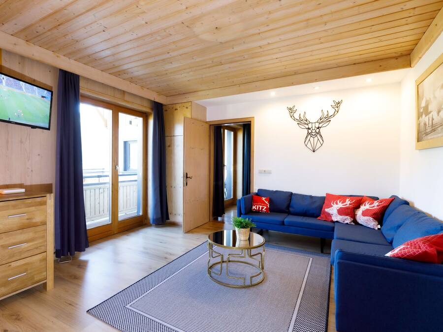A cozy living room with a wooden ceiling, a blue sofa with red cushions, a television mounted on the wall, and a deer head decoration.