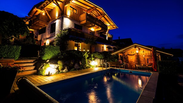 A large, multi-story wooden house with a swimming pool, surrounded by landscaped gardens and illuminated by warm lighting. - Landhaus Kitzblick (Piesendorf)