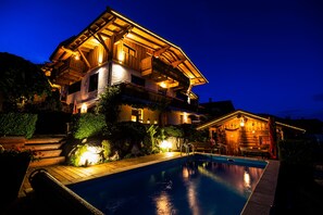 Una casa grande de madera, de varios pisos, con piscina, rodeada de jardines ajardinados e iluminada con luces cálidas.