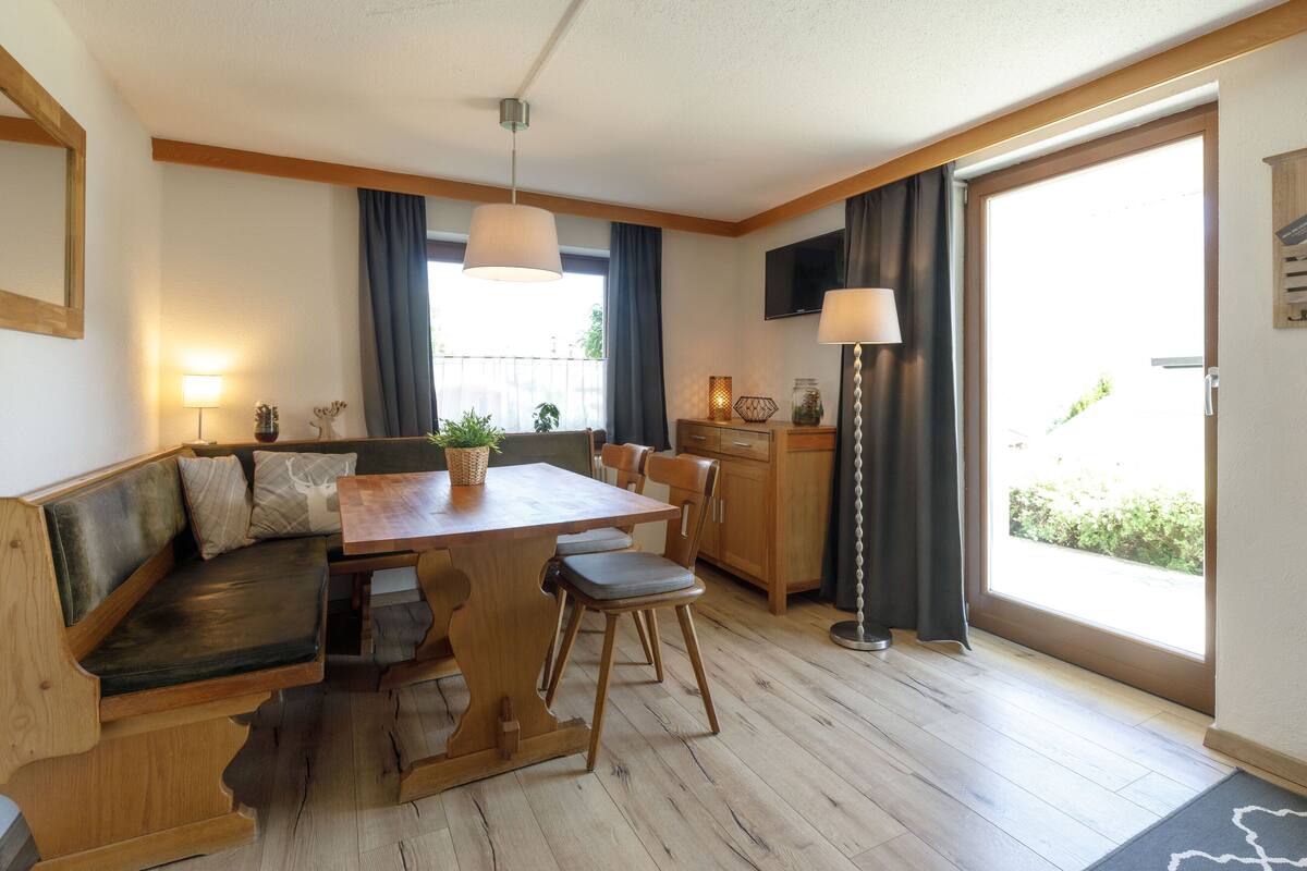A cozy room with a wooden bench, a dining table, and chairs. There is a TV mounted on the wall and a window with curtains.