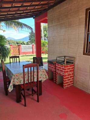 An outdoor patio with a brick barbecue grill, a table with chairs, and a view of a mountain range. - Newly renovated house with barbecue, 300 meters from Cordeirinho beach. (Maricá)