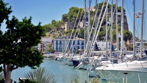 A marina with multiple sailboats docked, a hotel named "Hotel Eros" in the background, and a rocky hill with trees.