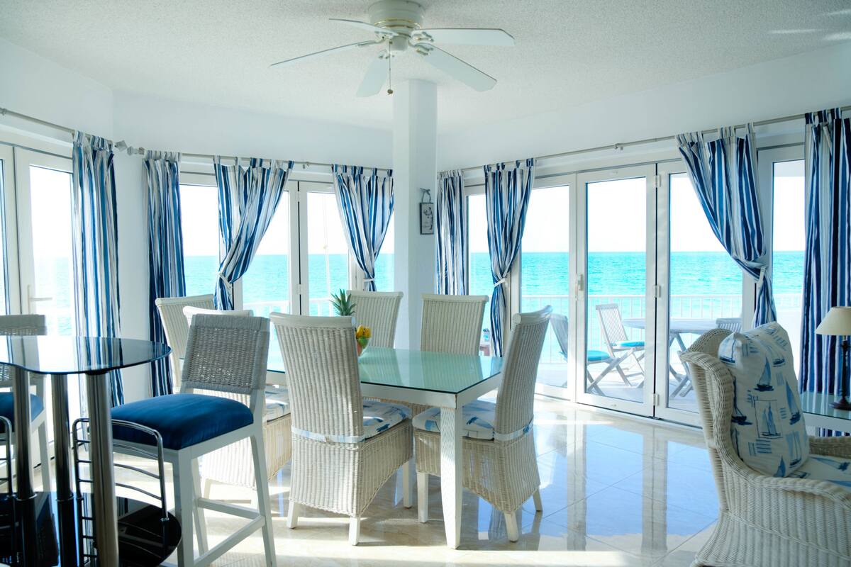 A dining area with a glass table and white wicker chairs, large windows with blue curtains, and a view of the ocean.