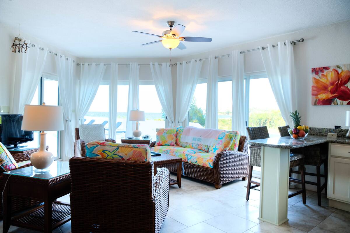 A bright living room with wicker furniture, a ceiling fan, and a kitchen area with a fruit bowl.
