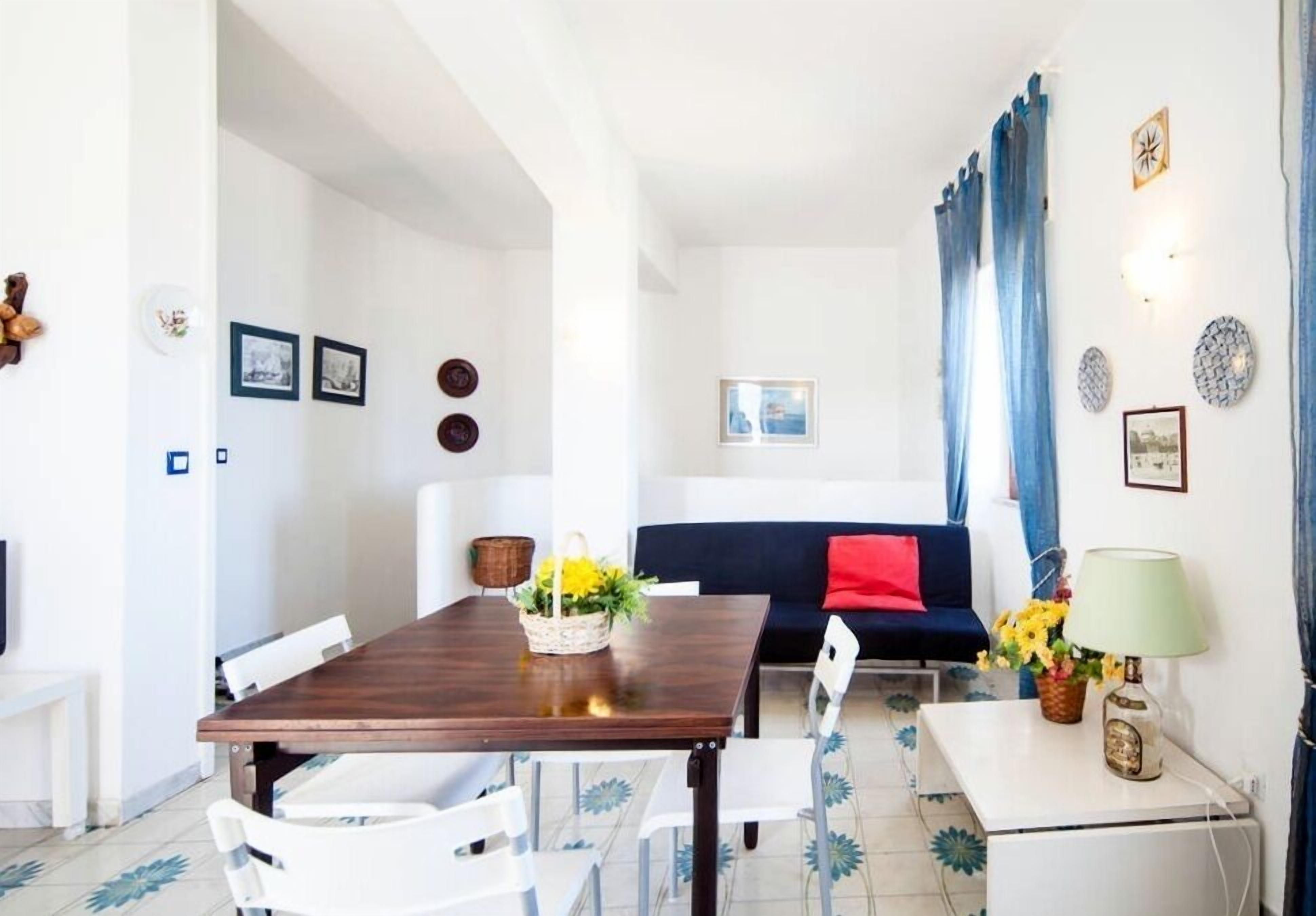 A dining area with a wooden table and white chairs, a sofa with a red cushion, and a lamp on a side table.