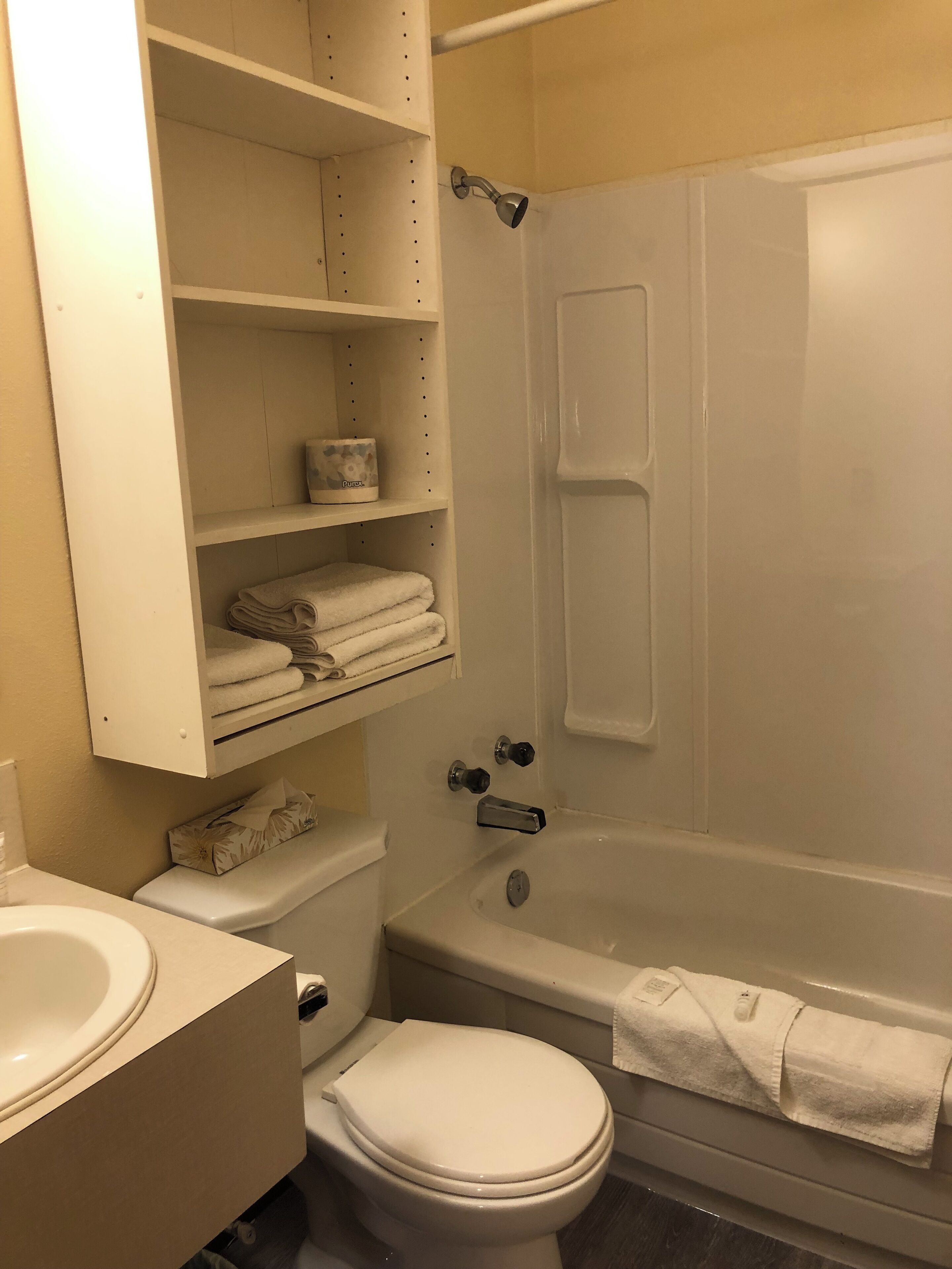 Un baño con inodoro, lavamanos y bañera. Hay repisas sobre el inodoro con toallas dobladas y un artículo de tocador.