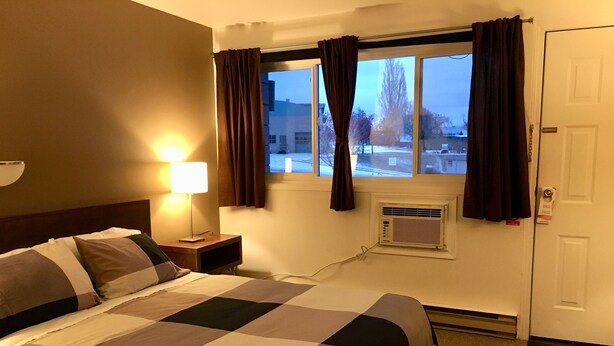 A hotel room with a bed, a nightstand, a lamp, a window with curtains, an air conditioning unit, and a door with a lock.