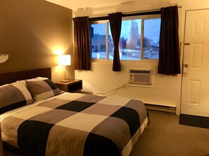 A hotel room with a bed, a nightstand, a lamp, a window with curtains, an air conditioning unit, and a door with a lock. - The Canterbury Inn of Downtown Invermere (Invermere)