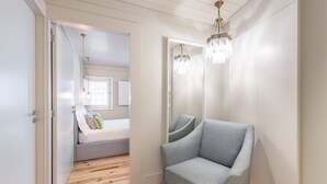 Ein Flur mit einem grauen Sessel, Holzboden und einem Spiegel, der ein Bett mit Kissen reflektiert.