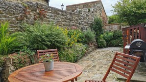 A stone patio with a round wooden table and two chairs, surrounded by lush greenery and a stone wall.