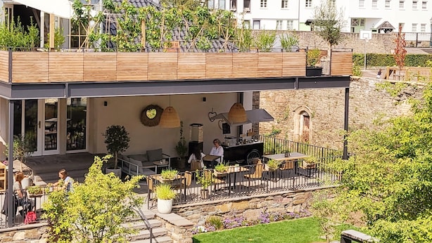 Un edificio moderno con una zona de estar al aire libre cubierta, muros de piedra y vistas a una colina con árboles y casas.