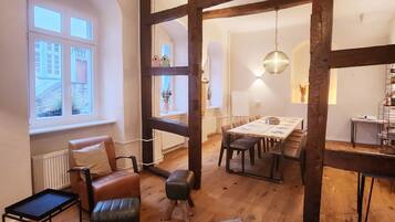 Una habitación amplia con vigas de madera a la vista, un comedor con una mesa larga y una zona de estar acogedora con un sillón y reposapiés de cuero.