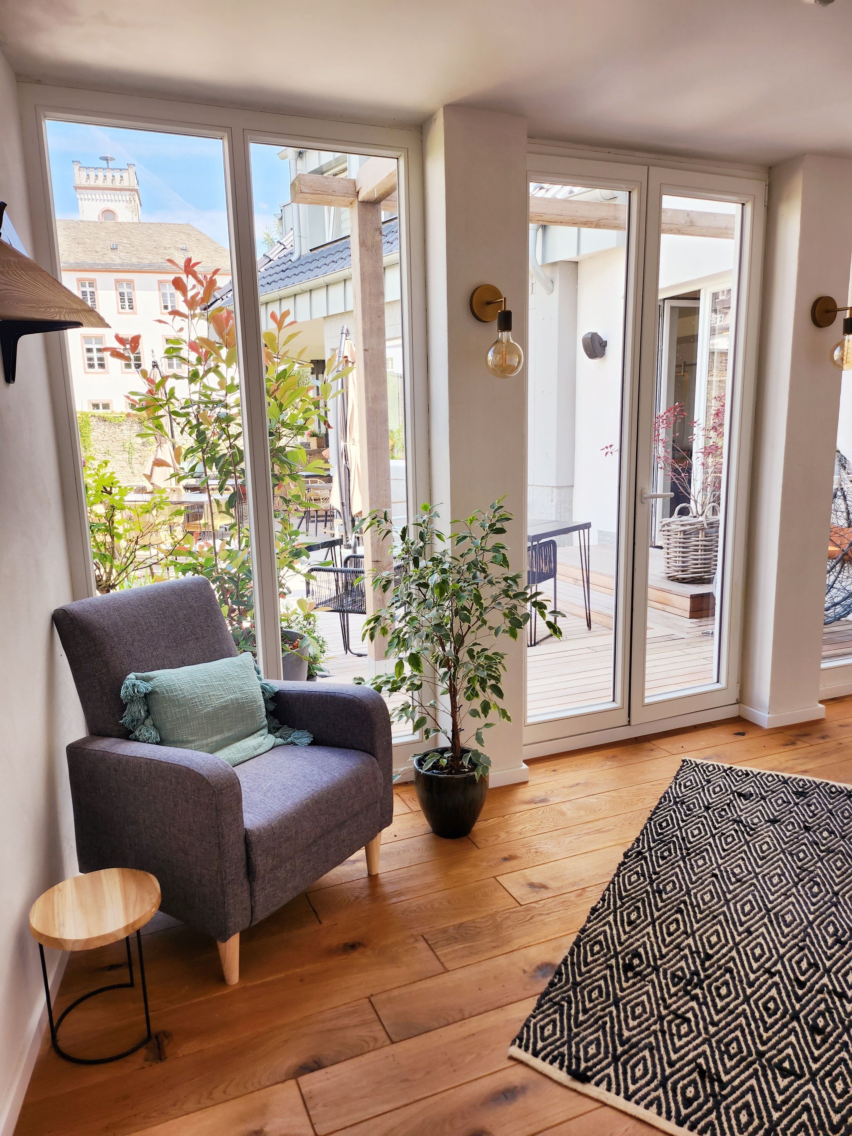 Ein gemütliches Wohnzimmer mit einem grauen Sessel, einem kleinen Holztisch, einem Blumentopf und einem gemusterten Teppich.