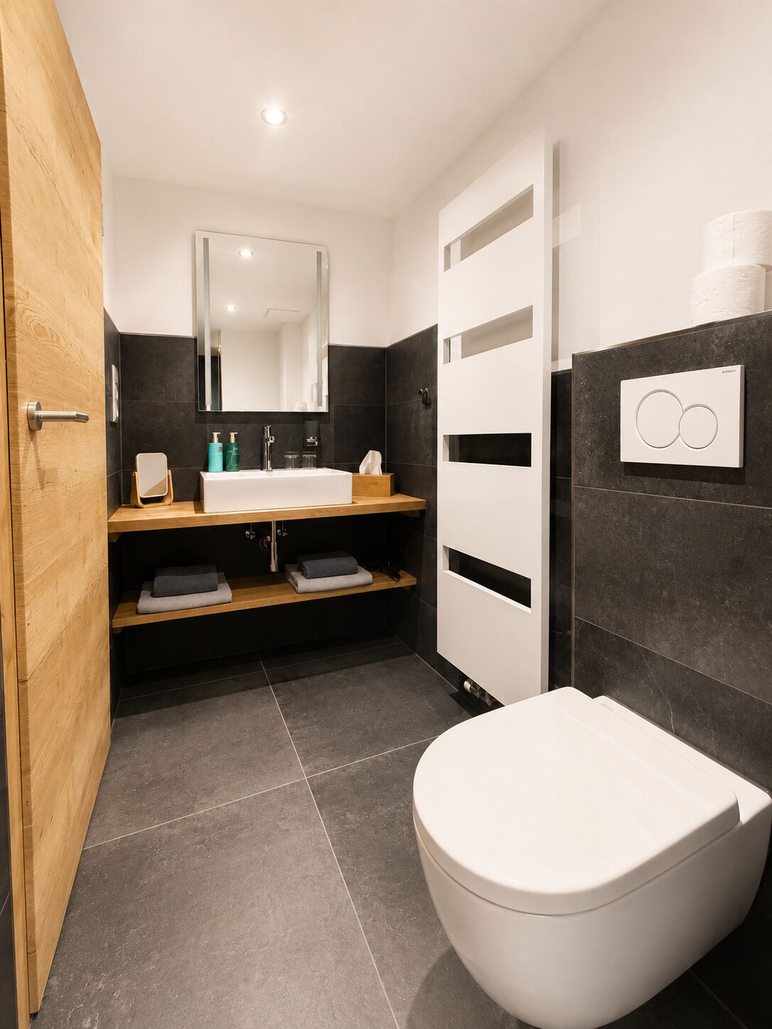 A modern bathroom with a white toilet, dark tiled walls, a wooden vanity, and a large mirror.