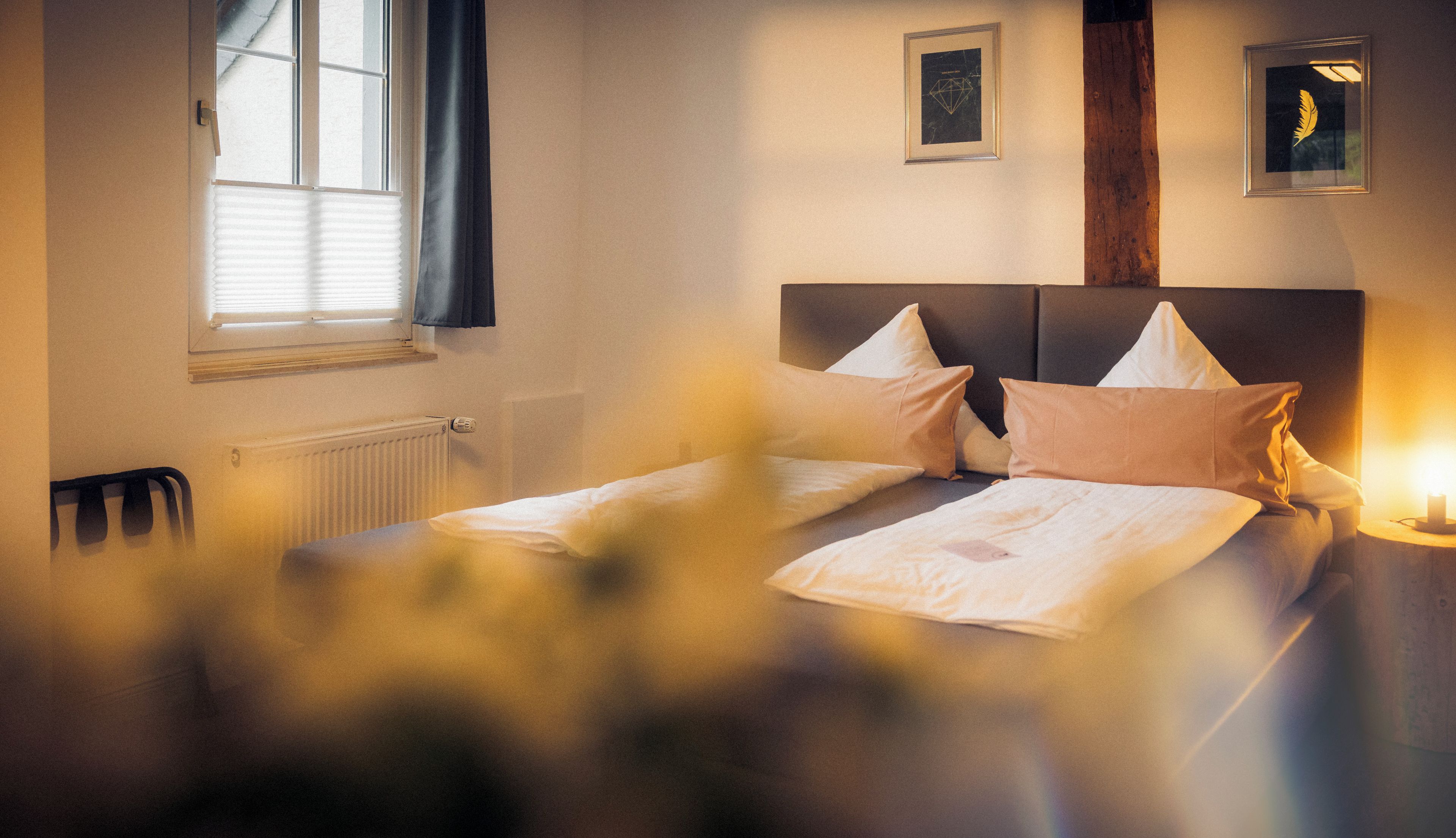 A hotel room with a bed, a radiator, a window with blinds, and two framed pictures on the wall.