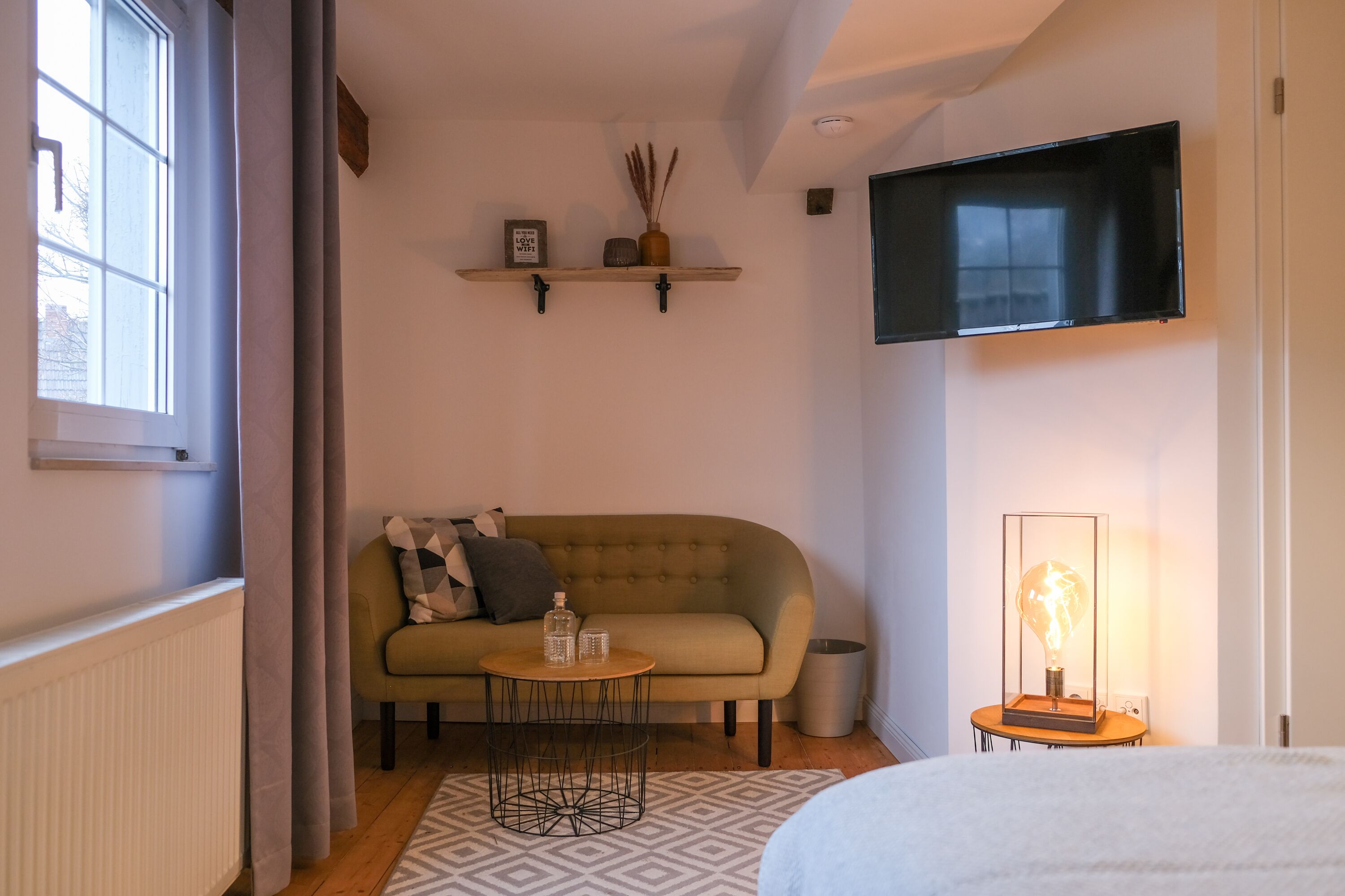 Una habitación acogedora con sofá, una mesita redonda, televisor montado en la pared y una lámpara decorativa.
