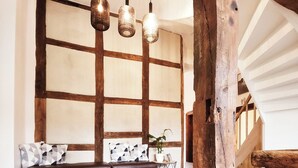 Un interior rústico con vigas de madera a la vista, suelo de baldosas de piedra, un banco de madera con cojines e iluminación colgante.
