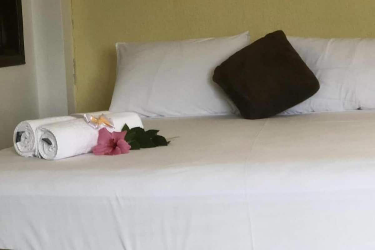 Una cama bien tendida con sábanas blancas, una toalla blanca enrollada, una flor rosa y una almohada marrón.