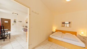 A hotel room with a bed, bedside lamps, a wooden headboard, a small bedside table, and a view of the dining area. - Apartment No. 14 in Vienna near Schönbrunn/Westbahnhof | Keyless Check-In (Rudolfsheim-Fünfhaus)