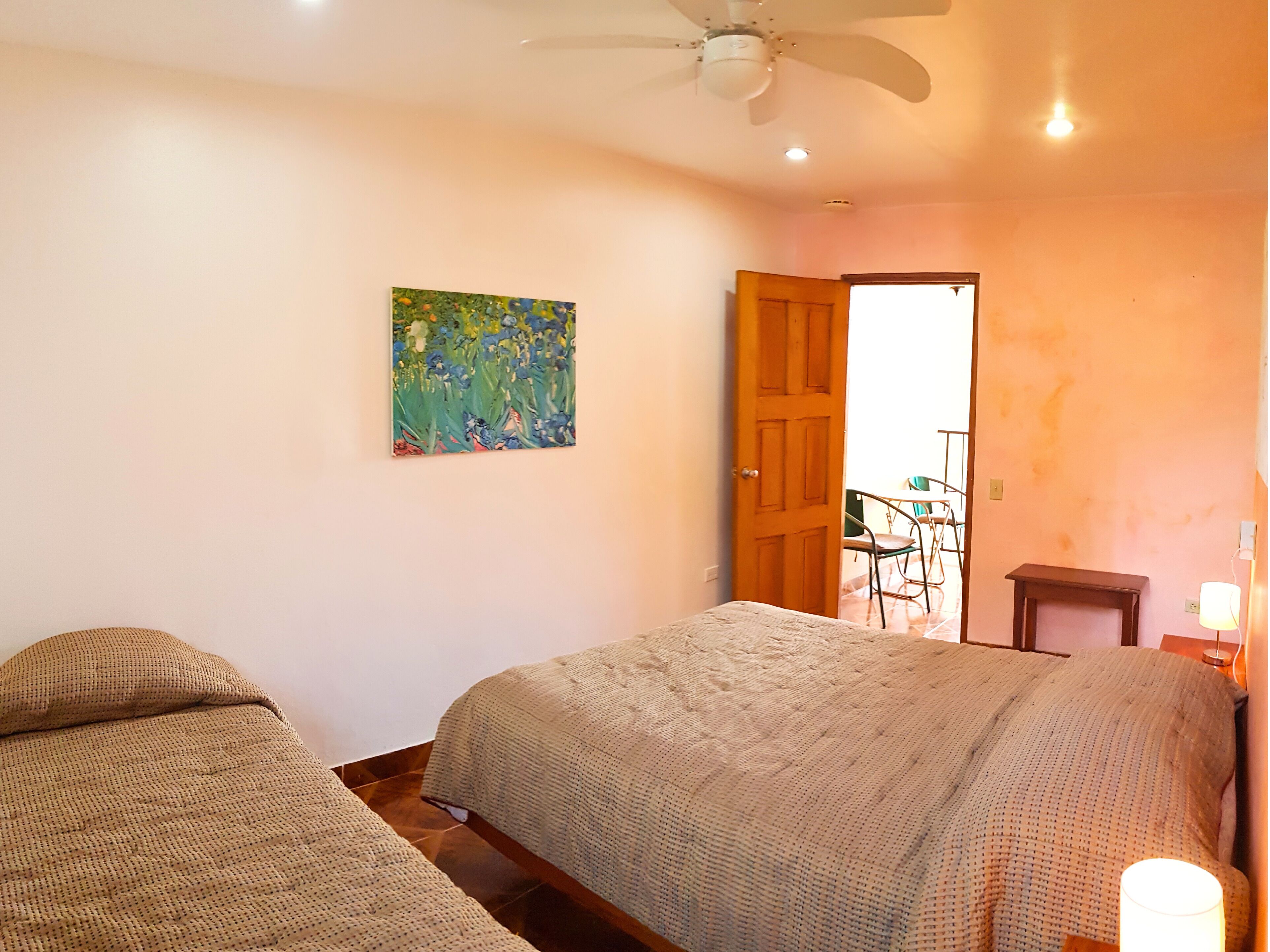 A bedroom with two beds, a ceiling fan, a painting, and a door leading to a balcony.