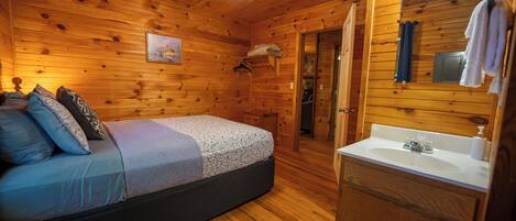 Un dormitorio de cabaña de madera con una cama, un ventilador de techo, un baño con lavamanos y espejo, y una puerta que da a otra habitación.