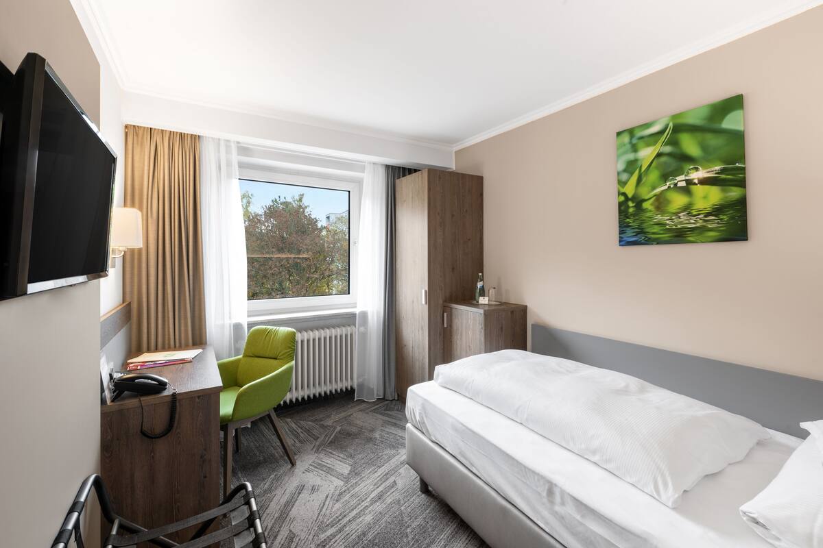 Habitación de hotel con cama, escritorio con silla, televisor, radiador y ventana con cortinas.