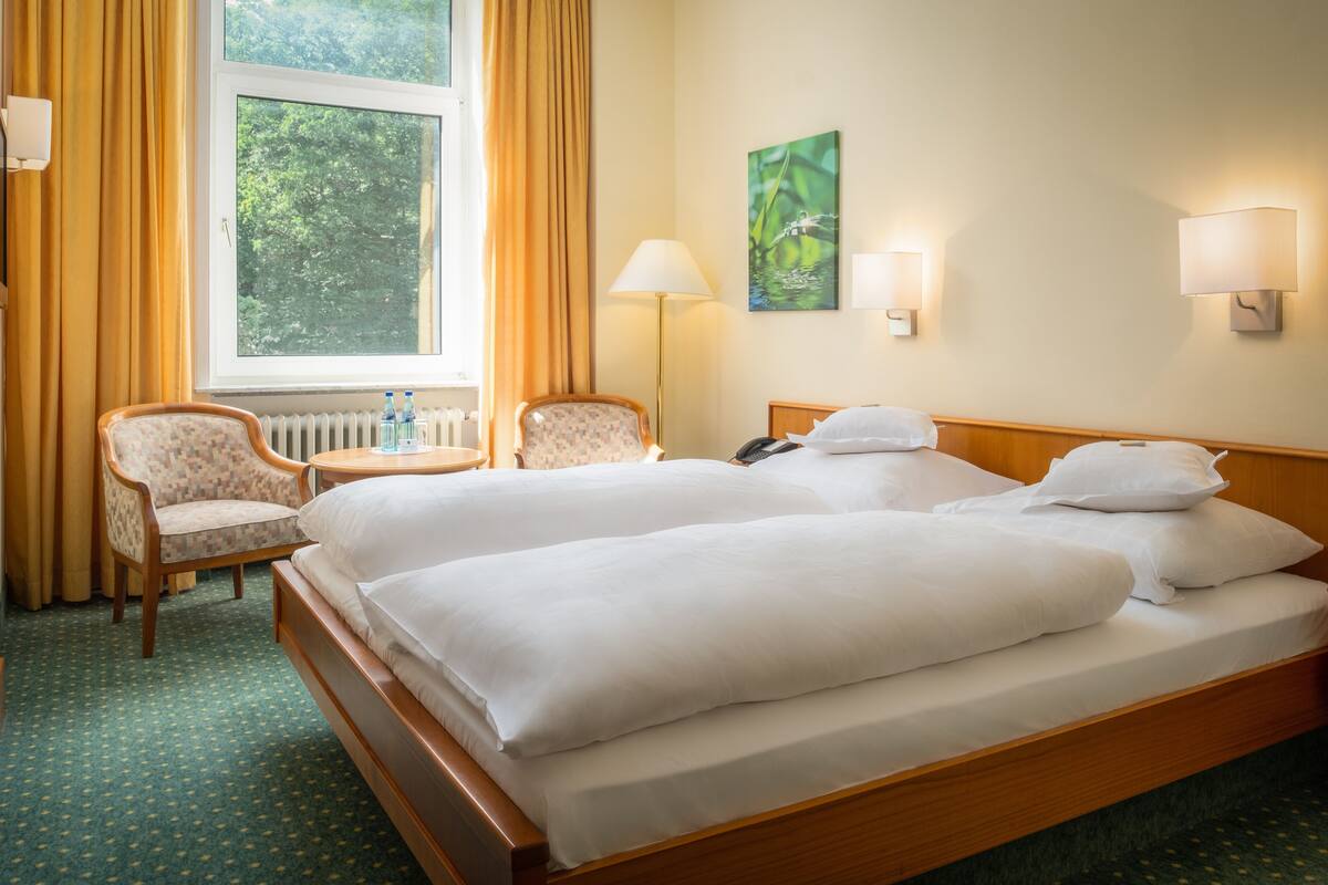 Habitación de hotel con dos camas, una silla, una lámpara y un ventanal con cortinas.