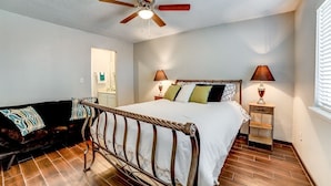 A bedroom with a bed, a ceiling fan, a nightstand with a lamp, a wooden floor, and a dark-colored sofa. - Stay in a classically designed apt near ORU, exercise trails, and restaurants! (Tulsa)