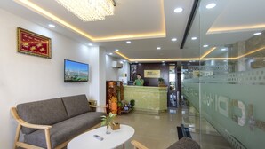A hotel reception area with a sofa, a reception desk, a television, and a chandelier. - Hodi Hotel (Da Nang)
