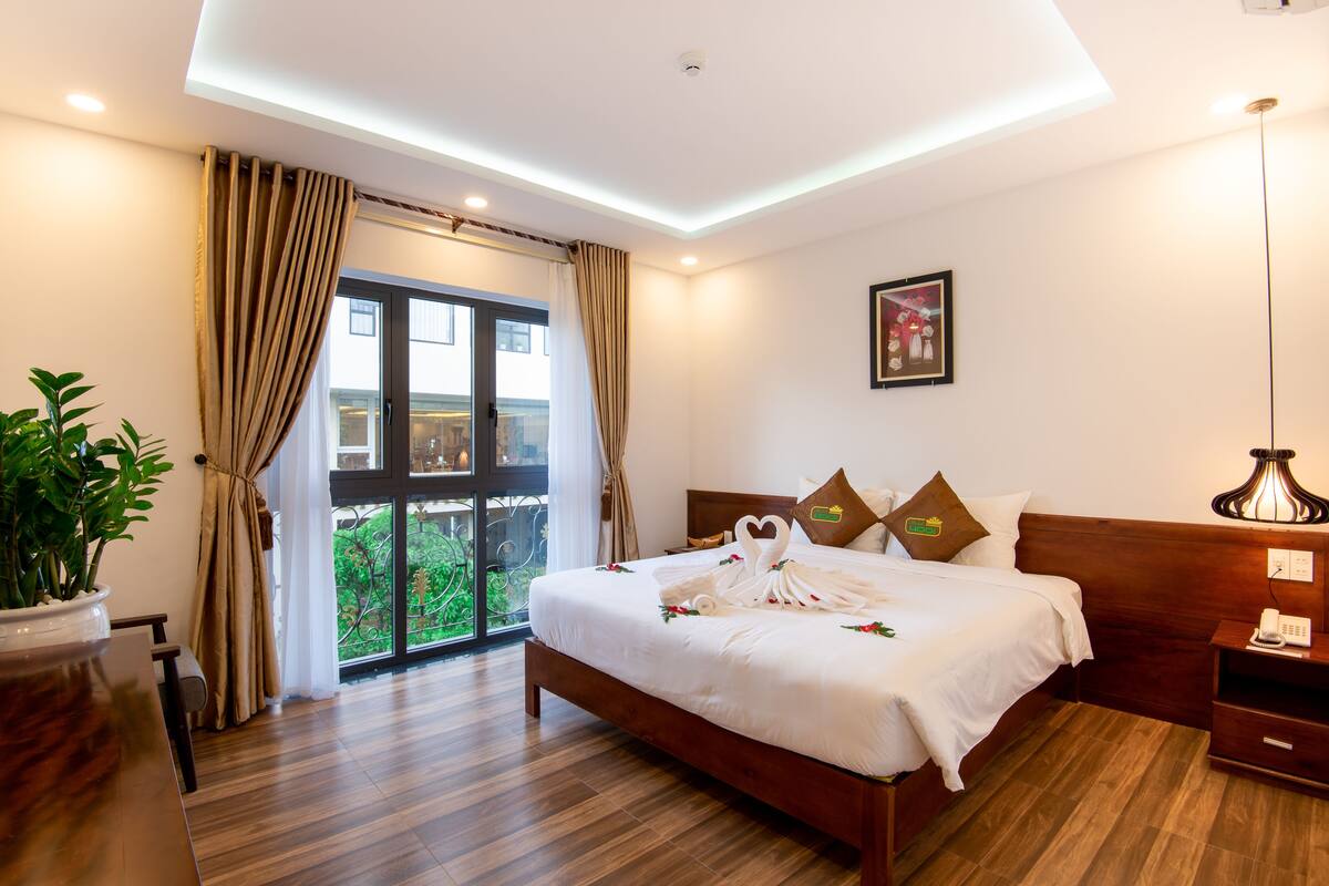 Habitación de hotel con una cama grande, suelo de madera, una planta en maceta, vistas a través de una puerta de cristal y una mesita de noche con lámpara.