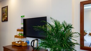 A hotel room with a flat-screen TV, a wooden coffee table with a kettle, a potted plant, and a wall-mounted shelf with toiletries. - Hodi Hotel (Da Nang)