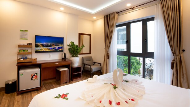 A hotel room with a bed, a desk, a television, a small refrigerator, a potted plant, and a window with curtains. - Hodi Hotel (Da Nang)