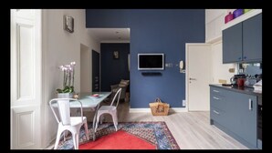 Een moderne keuken met blauwe kastjes, een eethoek met een rode vloerkleed, en een televisie aan de muur.