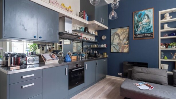 Een moderne keuken met grijze kasten, een ingebouwde oven en een aanrecht met diverse apparaten. In de woonkamer staat een grijze bank en een boekenkast met decoratieve objecten.