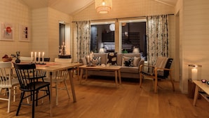 A cozy, well-lit room with wooden floors and walls, featuring a dining table with chairs, a sofa, and a small table with candles. - Fjällstugan (Bruksvallarna)