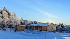 A wooden cabin with a snow-covered roof, surrounded by a forest and a mountain in the background. - Fjällstugan (Bruksvallarna)