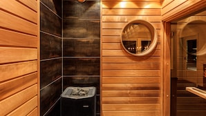 A sauna room with wooden walls, a circular mirror, and a black heater with rocks. - Fjällstugan (Bruksvallarna)