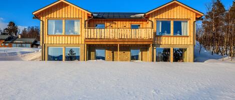 A two-story wooden cabin with large windows and a balcony, surrounded by snow.