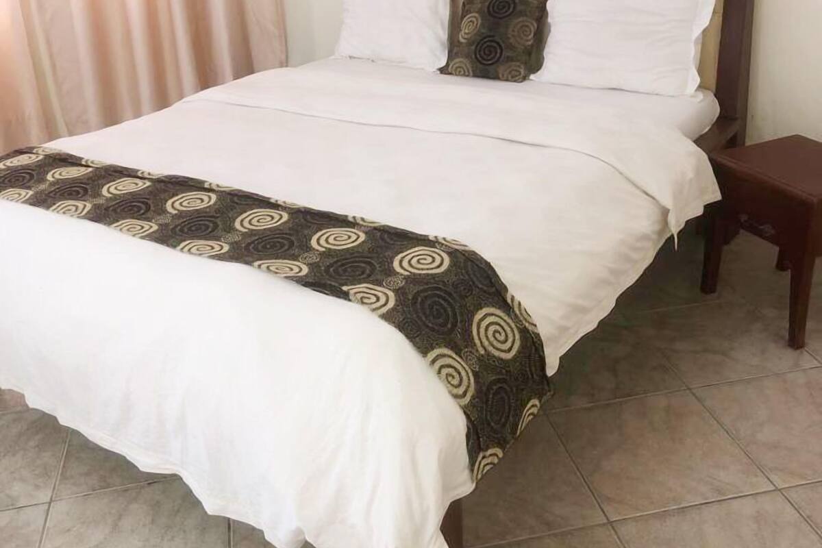 A neatly made bed with a patterned bedspread and two pillows, a wooden nightstand beside it, and a window with curtains.