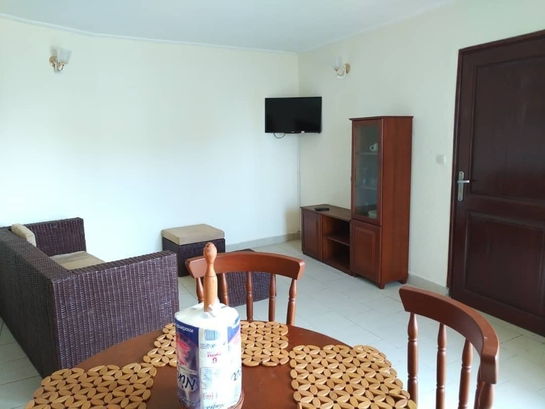 A living room with a sofa, a dining table with a can of milk, a wooden cabinet, and a TV mounted on the wall.