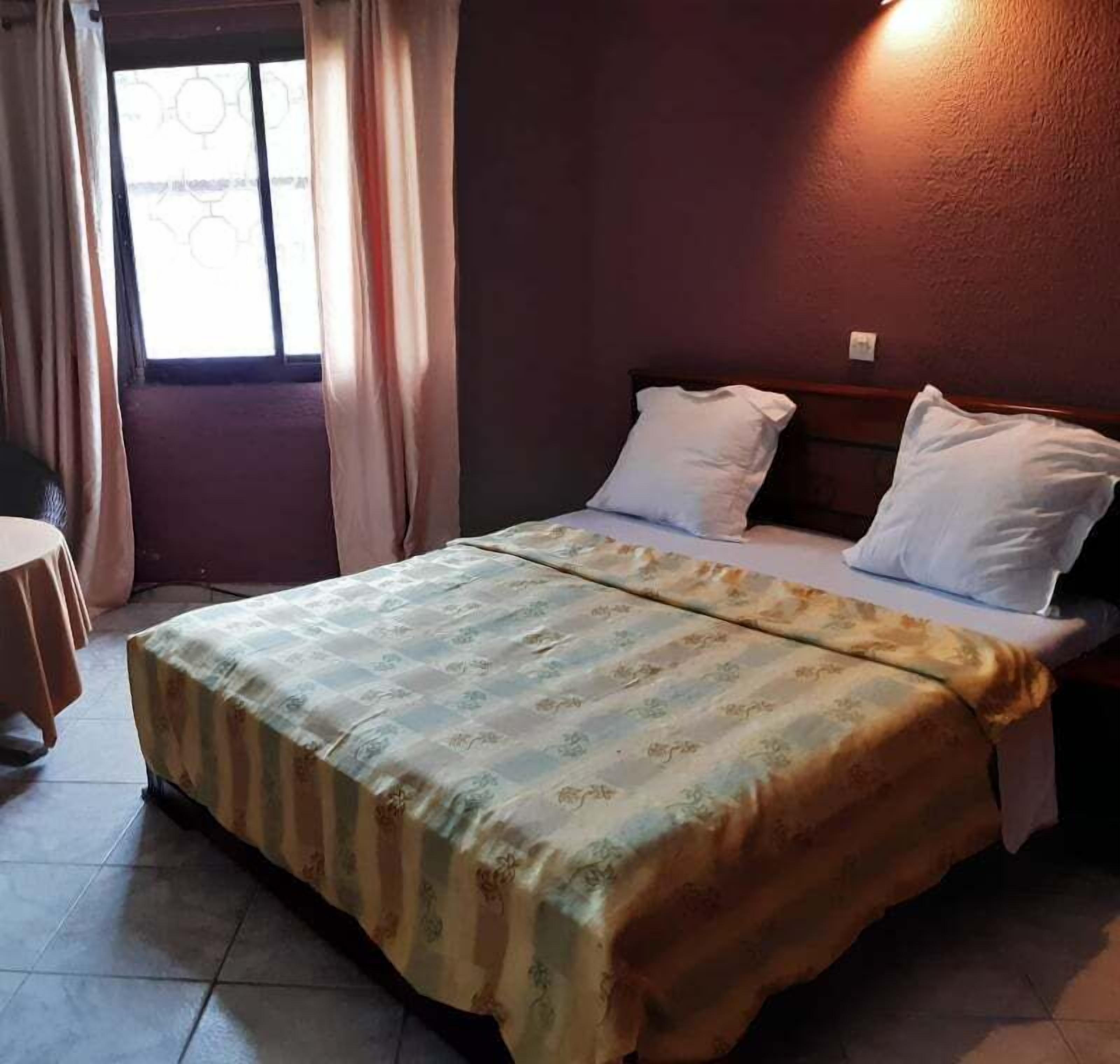 A hotel room with a bed, two pillows, a bedside table, and a window with curtains.