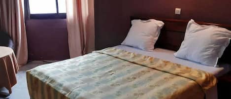 A hotel room with a bed, two pillows, a bedside table, and a window with curtains.