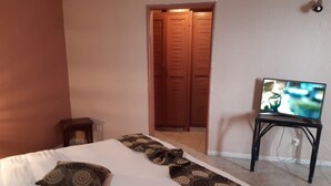 A hotel room with a bed, a television on a stand, a door, and a small table. - Hôtel Les Polygones (Kribi)