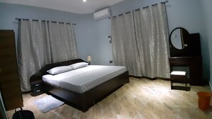 Un dormitorio con cama, cortinas, ventilador de techo, unidad de aire acondicionado, espejo y un cubo de basura pequeño.